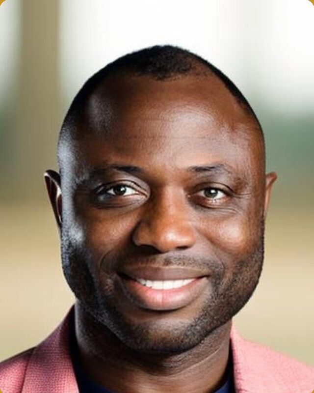 A portrait of a smiling man in a pink jacket over a navy shirt, against a warm blurred background.