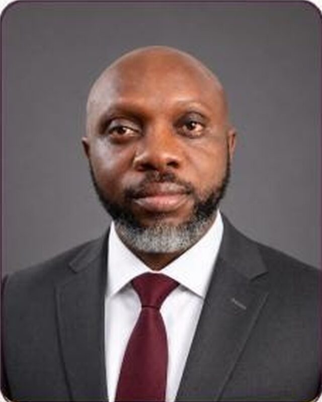 A studio portrait of a bald man with a grey beard, in a charcoal suit, white shirt and maroon tie, against a dark grey background.