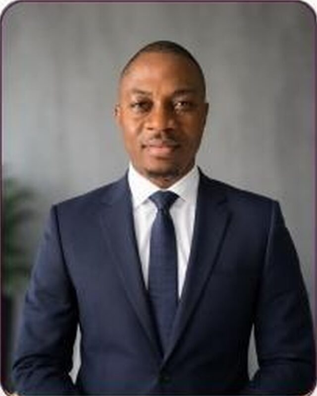 A portrait of a man in a navy suit, white shirt and navy tie, against a grey wall with a plant at frame left.
